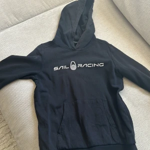 Svart hoodie från Sail Racing - Säljer en svart hoodie från Sail Racing med vit logga på bröstet. Den har en klassisk känguruficka och en bekväm huva. Perfekt för en avslappnad stil. Hör av dig vid intresse!