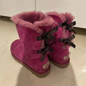 Rosa boots från UGG med rosetter - Säljer ett par rosa UGG boots med mysig pälsfoder och dekorativa svarta rosetter på baksidan. Perfekta för att hålla fötterna varma och stiliga under kalla dagar. Dem är nästan som nya. 
