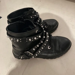 Svarta boots med nitar - Snygga svarta boots med nitar och spännen. De har en robust design med snörning och dragkedja för enkel på- och avtagning. Perfekta för en edgy stil.