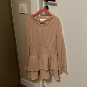 Beige klänning från Gina Tricot - Säljer en söt beige klänning från Gina Tricot med volangdetaljer och långärmad design. Klänningen har en smickrande passform med elastisk midja och ett diskret prickigt mönster. Perfekt för både vardag och speciella tillfällen.