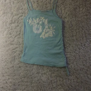 Turkos linnetopp från KappAhl - Säljer en söt turkos linnetopp från KappAhl med blommigt tryck och siffran 61 på framsidan. Toppen har smala axelband och en avslappnad passform. Perfekt för varma sommardagar! 🌸(sorry om det är skrynkligt äger inget stryckjärn)