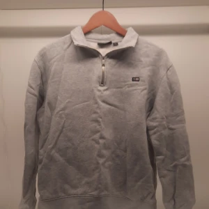 Grå tröja med dragkedja från Lexington - Säljer en stilren grå tröja från Lexington med dragkedja och hög krage en liten orange fläck bredvid Lemingtonmärket men den går nog att få bort om man har rätt tvättmedel. Perfekt för en avslappnad look. Tröjan har en liten Lexington-logga på bröstet och är tillverkad i ett mjukt material.