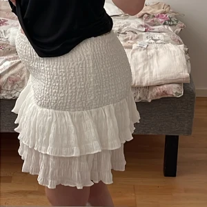 Vit volangkjol - Säljer en söt vit volangkjol med elastisk midja. Kjolen har flera lager av volanger som ger en lekfull look. Perfekt för varma dagar och kan enkelt matchas med olika toppar.