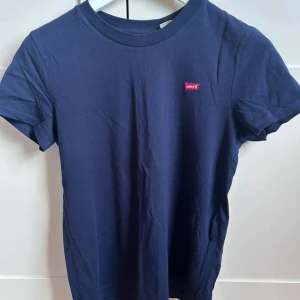 Mörkblå t-shirt från Levi's - Säljer en stilren mörkblå t-shirt från Levi's med en liten röd logga på bröstet. Perfekt för en casual look. T-shirten är kortärmad och tillverkad i mjukt material.