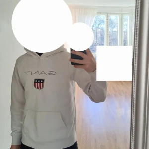 Vit hoodie från Gant - Säljer en stilren vit hoodie från Gant med deras ikoniska logga och ett sköldmärke på bröstet. Tröjan har en klassisk känguruficka och långa ärmar. Perfekt för en avslappnad look.