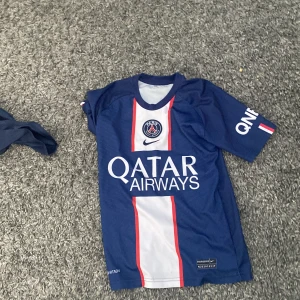 Blå PSG fotbollströja med Mbappé 7 - Säljer en blå PSG fotbollströja från Nike med Mbappé och nummer 7 på ryggen. Tröjan har korta ärmar och är tillverkad i Dri-FIT material för optimal komfort. Framsidan pryds av Qatar Airways logga och PSG-emblemet.