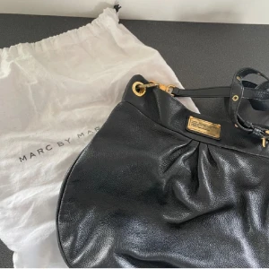 Svart axelväska från Marc by Marc Jacobs - Snygg svart axelväska i skinn från MARC BY MARC JACOBS med guldfärgade detaljer, tre innerfickor, varav en dragkedsförsedd. Axelrem och kort band finns till. Dustbag medföljer.  Bredd 29 cm, höjd 31 cm, djup ca 7 c  Skicka meddelande för fler bilder💞💞