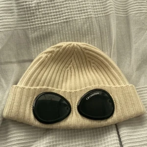 Gräd vit mössa med glasögondetalj från C.P. Company - Snygg gräd vit mössa från C.P. Company med unika glasögondetaljer framtill. Den är ribbad och tillverkad i ett mjukt material som ger en skön passform. Du får med ett armband 