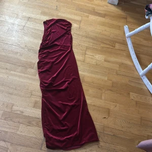 Vinröd långklänning - Elegant vinröd långklänning i ett mjukt och lyxigt material. Klänningen har en smickrande drapering och är axelbandslös, vilket ger en stilren och sofistikerad look. Perfekt för speciella tillfällen.