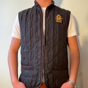 Svart quiltad väst från Morris - Säljer en snygg quiltad väst från Morris med dragkedja och broderad logga på bröstet. Västen har två praktiska fickor framtill och är perfekt inför våren!