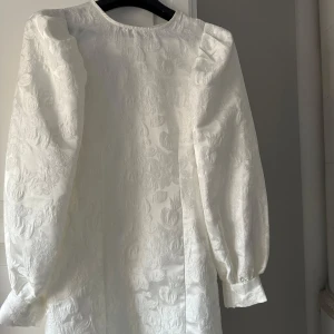 Vit klänning med blommigt mönster - Elegant vit klänning med ett subtilt blommigt mönster. Klänningen har långa ärmar och en klassisk rund halsringning. Perfekt för en stilren look.