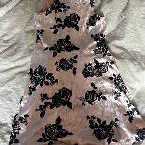 Rosa klänning med blommönster från Lindex - Säljer en söt rosa klänning från Lindex med svart blommönster. Klänningen har en ärmlös design och är gjord i ett glansigt material som ger en lyxig känsla. Perfekt för speciella tillfällen!
