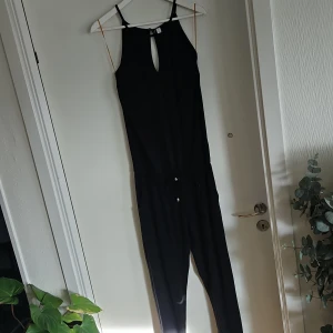 Svart ärmlös jumpsuit - Snygg svart ärmlös jumpsuit med smala axelband och en liten öppning framtill. Perfekt för en stilren look. Den har en knytning i midjan för en justerbar passform.