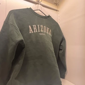 Grön sweatshirt med texten Arizona - Säljer en grön sweatshirt med texten 'Arizona U.S.A.' på bröstet. Tröjan har långa ärmar och en rund halsringning. Perfekt för en avslappnad stil.