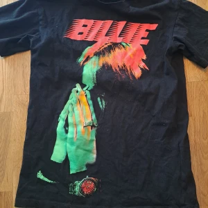 Svart t-shirt med tryck - Cool svart t-shirt med ett färgstarkt tryck av en person och texten 'BILLIE' i rött. Perfekt för fans av unik design och starka färger. T-shirten har korta ärmar och är gjord i ett bekvämt material.