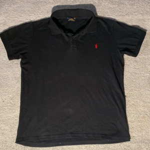 Svart pikétröja från Ralph Lauren - Säljer en klassisk svart pikétröja från Ralph Lauren med den ikoniska röda loggan på bröstet. Tröjan har korta ärmar och en stilren krage med knappar. Perfekt för en avslappnad men ändå stilfull look. Skickar inom 24h😋