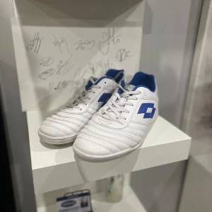 Vita och blå fotbollsskor från Lotto - Snygga vita fotbollsskor från Lotto med blå detaljer och snörning. Perfekta för fotbollsplanen med en stilren design och bekväm passform.