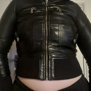 Svart skinnjacka med dragkedja - Snygg svart skinnjacka med dragkedja och ribbade muddar. Jackan har flera fickor med dragkedjor och knappar för en cool look. Perfekt för att ge din outfit en edgy touch.