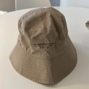 Beige bucket hat - Snygg beige bucket hat med diskret text på framsidan. Perfekt för en avslappnad stil och skydd mot solen. Tillverkad i ett slitstarkt material som ger en skön passform.