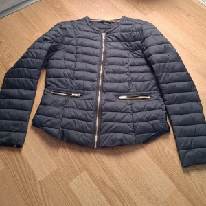 Mörkblå pufferjacka från Gina Tricot - Snygg mörkblå pufferjacka från Gina Tricot med dragkedja framtill och två dragkedjefickor. Jackan har en quiltad design och långa ärmar, perfekt för kyligare dagar.