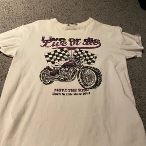 Vit t-shirt med motorcykeltryck från FB Sister - Cool vit t-shirt från FB Sister med ett stort tryck av en motorcykel och rutiga flaggor. Texten 'Live or die' och 'Move the soul' ger en edgy känsla. Perfekt för dig som gillar en rockig stil. Storleken är XS men skulle säga att den är mer S/M