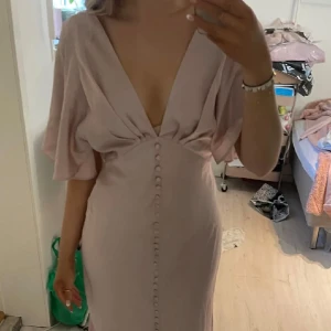 Balklänning  - Elegant ljusrosa klänning med djup v-ringning och knappar framtill. Klänningen har korta, vida ärmar och en figursydd passform som ger en smickrande siluett. Perfekt för speciella tillfällen. Med prislapp. Nypris 1069