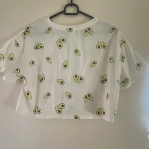 Vit croppad t-shirt med avokadotryck från Shein - Säljer en vit croppad t-shirt från Shein med ett lekfullt avokadotryck. T-shirten har korta ärmar och en avslappnad passform. Perfekt för en avslappnad stil!
