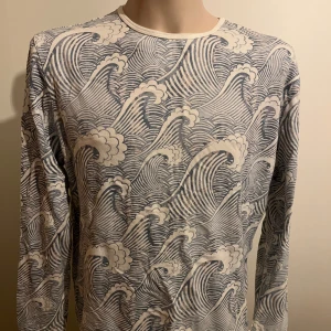 Vit och blå långärmad tröja med vågmönster från Homebound - Säljer en stilren vit och blå långärmad tröja från Homebound med ett unikt vågmönster. Perfekt för dig som vill ha något annorlunda i garderoben. Tröjan har en rund halsringning och är tillverkad i ett mjukt material.