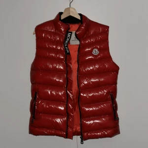 väst från Moncler, öppen för byten  - Säljer en snygg röd dunväst från Moncler i S. Västen är quiltad och har en glansig finish med dragkedja framtill. Perfekt för höst och vår när du vill hålla dig varm och stilren. Moncler-loggan syns tydligt på bröstet. lite problem i höger ficka