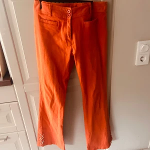 Lågmidjade byxor - Säljer ett par snygga orange lågmidjade byxor från Miss Fisher. De har en cool snörningsdetalj vid bensluten och är perfekta för att liva upp garderoben. Byxorna är i storlek 40 och har en bekväm passform.🧡🌞