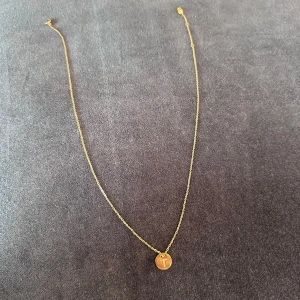 Guldfärgat halsband - Guldfärgat halsband med bokstavsberlock föreställande bokstaven T. Mycket gott skick. Kedjan går att justera till tre olika längder genom spännet.   Längd kedja: 47 cm Diameter berlock: 1 cm