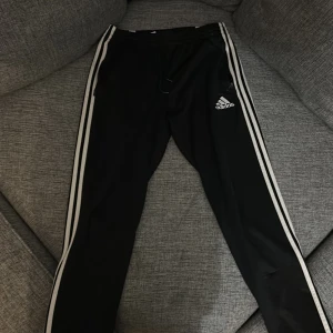 Svarta träningsbyxor från Adidas - Snygga svarta träningsbyxor från Adidas med de klassiska tre ränderna längs sidorna. Byxorna har en bekväm passform med snörning i midjan och är perfekta för träning eller en avslappnad stil.