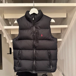 Svart dunväst från Polo Ralph Lauren - Storlek L i barnstorlek, 14-16 år. Säljer en snygg svart dunväst från Polo Ralph Lauren med dragkedja och broderad logga i rött på bröstet. Perfekt för kyligare dagar med sin quiltade design och höga krage.