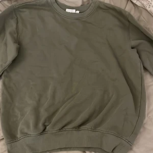 Olivgrön sweatshirt från KappAhl - Säljer en olivgrön sweatshirt från KappAhl i storlek 158/164. Tröjan har en klassisk rund halsringning och långa ärmar med ribbade muddar vid ärmslut och nederkant. Perfekt för en avslappnad stil.