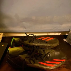 Svarta fotbollsskor från Adidas - Snygga svarta fotbollsskor från Adidas med röda detaljer och snörning. Perfekta för fotbollsplanen med en stilren design och bra grepp.