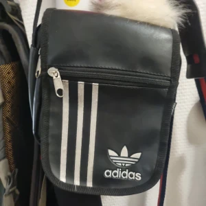 Svart axelväska från Adidas - Snygg svart axelväska från Adidas med klassiska vita ränder och logotyp. Väskan har en praktisk dragkedja och är perfekt för att bära med sig det nödvändigaste. Kompakt och stilren design som passar till många tillfällen.