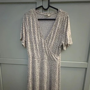 Beige blommig klänning - Säljer en söt beige klänning från Indiska med blommigt mönster. Klänningen har en omlott-design med v-ringning och korta ärmar.