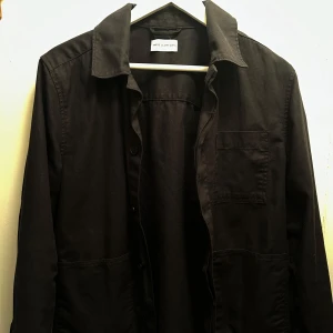 Svart overshirt från These Glory Days - Snygg svart overshirt från Some Glory Days med klassisk krage och knappar framtill. Perfekt för lager-på-lager-stil. Har en bröstficka för extra stil och funktion.
