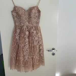 Beige spetsklänning med blommigt mönster - Elegant beige klänning med vacker spets och blommigt mönster. Axelbandslös design med detaljerade broderier och pärlor. Perfekt för speciella tillfällen.