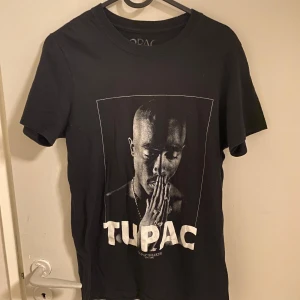 Svart T-shirt med Tupac-tryck - Cool svart T-shirt med ett stort tryck av Tupac på framsidan. Perfekt för fans av hiphop och streetwear. T-shirten har en klassisk rund hals och korta ärmar.