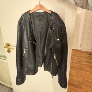 Svart skinnjacka med dragkedja - Snygg svart skinnjacka med asymmetrisk dragkedja och silverfärgade detaljer. Jackan har långa ärmar och två framfickor med dragkedjor. Perfekt för en cool och stilren look.