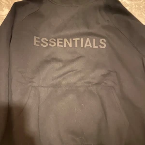 Svart Essentials hoodie från Fear of God - Säljer en svart Essentials hoodie från Fear of God. Den har en stor logga på bröstet och en mindre på ärmen. Perfekt för en stilren och avslappnad look.