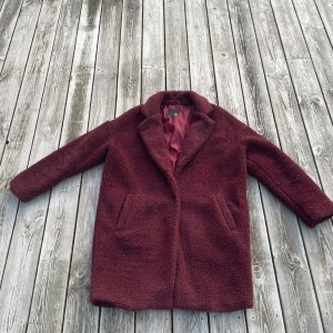 Vinröd teddyjacka - Mysig vinröd teddyjacka med långa ärmar och två framfickor. Perfekt för kyliga dagar med sin mjuka och varma textur. Fodrad insida för extra komfort.