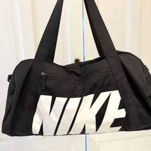 Svart sportväska från Nike - Säljer en svart sportväska från Nike med stort vitt logotryck. Väskan har dubbla handtag och en justerbar axelrem för bekväm bärning. Den har en dragkedja upptill och flera fack för enkel organisering. Perfekt för träning eller resor. Mått: 56cm lång, 24cm bred, 29cm hög.
