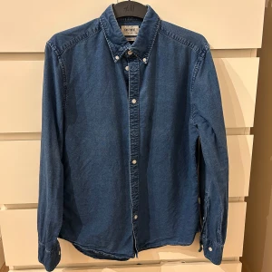 Blå jeansskjorta från Gant - Snygg blå jeansskjorta från Brothers med klassisk krage och knappar framtill. Skjortan har långa ärmar och en stilren design som passar perfekt för en avslappnad look. Den är använd ett fåtal gånger men är för liten i storleken för mig.