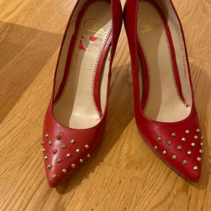 Röda pumps med nitar - Snygga röda pumps med spetsig tå och klack. Skorna är dekorerade med guldnitar framtill, vilket ger dem en edgy look. Perfekta för att sätta en färgklick på din outfit.