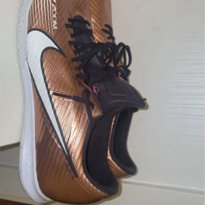 Bronsfärgade Nike Zoom sneakers - Snygga bronsfärgade Nike Zoom sneakers med vit sula och svart snörning. Perfekta för en sportig look med en modern touch. Den ikoniska Nike-loggan i vitt syns tydligt på sidan.