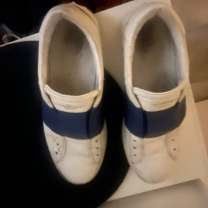 Vita sneakers med blå detalj - Snygga vita sneakers med en bred blå rem över vristen. Perfekta för en stilren och modern look. Skorna har en klassisk design med rund tå och är tillverkade i syntetmaterial.