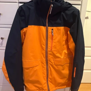 Orange och marinblå vinterjacka - Snygg och funktionell orange och marinblå vinterjacka med huva och dragkedja. Jackan har praktiska fickor med dragkedjor.