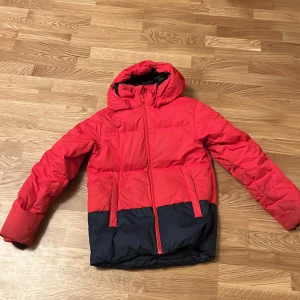 Röd och marinblå dunjacka med huva - Snygg och varm röd dunjacka från Reima med marinblå detaljer. Jackan har en praktisk huva och dragkedja framtill. Perfekt för kyliga dagar.